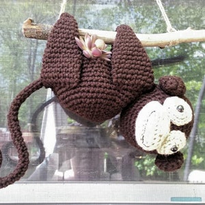 May include: A brown crocheted monkey hanging upside down from a branch. The monkey has a long tail and a small plant growing out of its back.