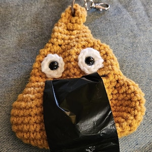 May include: A crocheted poop emoji keychain with googly eyes and a black plastic bag attached. The keychain is brown and has a silver metal ring.