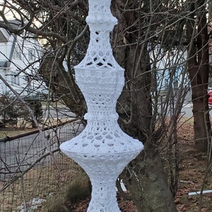 Op de afbeelding: Een witte gehaakte macramé hanglamp met een franje. De lamp hangt aan een tak.