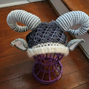 May include: A handmade crochet hat with large, curved horns in shades of gray and white. The hat features a dark gray open-weave crown, white trim, and small ear details. The hat is displayed on a purple wire stand.