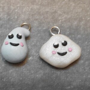 Op de afbeelding: Twee kleine, witte, klei-charms met geschilderde gezichten. Een charme is gevormd als een geest en de andere is een vierkant. Beide charms hebben zwarte ogen, zwarte glimlachen en roze wangen.