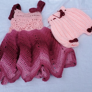 Puede incluir: Un conjunto de vestido y bombachos de bebé de ganchillo en tonos rosa y burdeos. El vestido tiene un dobladillo festoneado y los bombachos tienen un detalle de lazo.