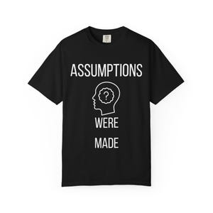 Könnte beinhalten: Ein schwarzes T-Shirt mit kurzen Ärmeln und dem weißen Text ASSUMPTIONS WERE MADE. Zwischen den Wörtern befindet sich eine weiße Strichgrafik eines menschlichen Kopfprofils mit einem Fragezeichen in einer wolkenartigen Form.