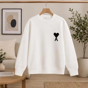 Könnte beinhalten: Ein weißes Langarm-Sweatshirt mit Rundhalsausschnitt und einem schwarzen, gestickten Herz- und A-Logo auf der linken Brust. Das Kleidungsstück verfügt über einen gerippten Kragen, Bündchen und Saum sowie ein kleines weißes Markenetikett im Nackenbereich.