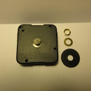 May include: A black square quartz clock movement mechanism with a central gold-colored metal shaft. Included are a gold-colored hexagonal nut, a thin gold-colored metal washer, and a circular black rubber gasket, all arranged to the right side.