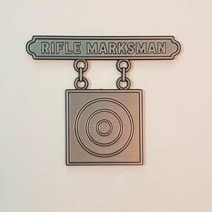 May include: A silver-toned metal badge featuring a rectangular bar at the top with the text RIFLE MARKSMAN embossed in black. Two small circular links connect the bar to a square pendant below, which displays a centered target graphic with three rings.