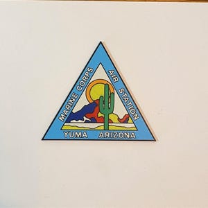 May include: A triangular enamel pin featuring a light blue border with the text MARINE CORPS AIR STATION YUMA ARIZONA. The center depicts a desert landscape with a yellow sun, a green saguaro cactus, and colorful abstract mountains.