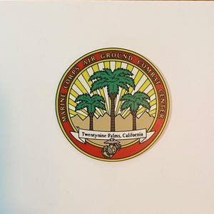 May include: A circular sticker featuring the Marine Corps Air Ground Combat Center emblem. It displays three green palm trees against a yellow sunburst and tan mountains, with a red border and the text Twentynine Palms, California at the base.