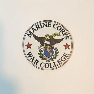 May include: A circular sticker featuring the Marine Corps War College emblem. The design includes a central eagle, globe, and anchor, with the text 'MARINE CORPS WAR COLLEGE' in black lettering around the border and two red stars on the sides.