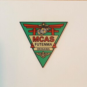 May include: A triangular patch featuring a green background with a red torii gate, the United States Marine Corps emblem, and the text MCAS FUTENMA OKINAWA in black and red lettering, with a small gold winged emblem at the bottom point of the triangle.