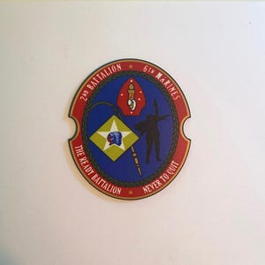 May include: A circular emblem for the 2nd Battalion 6th Marines featuring a red border with gold text reading 2ND BATTALION 6TH MARINES and THE READY BATTALION NEVER TO QUIT. The center displays a blue field with a torch, a flag, and a silhouette.