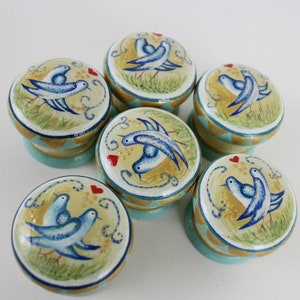 Puede incluir: Juego de seis pomos decorativos para armarios. Cada pomo presenta un diseño pintado a mano de pájaros azules, corazones rojos y detalles dorados sobre un fondo amarillo. Los pomos tienen una base azul claro.