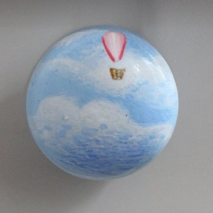 May include: A round, blue and white ceramic drawer knob with a painted hot air balloon in the sky. The balloon is pink and white with a gold basket.