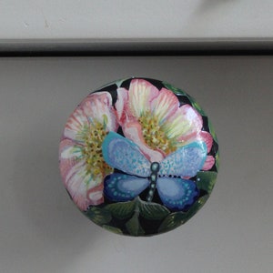 May include: A round cabinet knob with a painted butterfly and floral design. The knob is mounted on a white drawer. Pink flowers are in the background.
