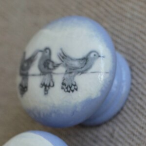 May include: Three pale blue cabinet knobs with a white top featuring a black and grey bird design. The knobs are arranged vertically, showcasing the bird illustrations on each knob. The background is a neutral fabric.