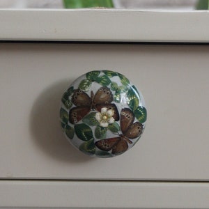 May include: A round drawer knob with a butterfly motif. The knob displays brown butterflies, green leaves, and small white flowers against a light backdrop. It is fixed to a light grey drawer.
