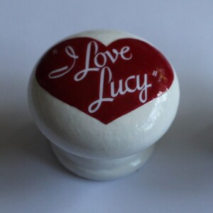 Op de afbeelding: Twee witte kastknoppen met een rood hartontwerp. Het hart bevat de tekst "I Love Lucy" in witte letters. De knoppen zijn rond met een kleine basis.