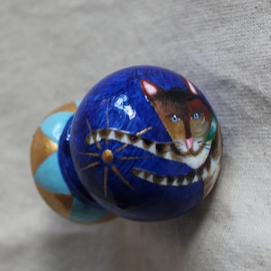 Puede incluir: Un pomo de madera pintado de azul y dorado con una cara de gato pintada en la parte superior. El gato tiene pelaje marrón y blanco y ojos azules.