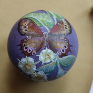 May include: A purple painted sphere with a brown and white butterfly, white flowers, and green leaves painted on it.