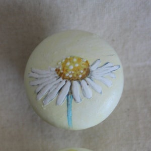 Könnte beinhalten: Ein runder, hellgelber dekorativer Knopf mit einem handgemalten Gänseblümchen-Design. Die Gänseblümchen haben weiße Blütenblätter, eine gelbe Mitte mit weißen Punkten und einen hellblauen Stiel. Der Knopf ist wahrscheinlich aus Holz.