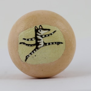 Może przedstawiać: Ozdobny przedmiot w kształcie jaja, w kolorze beżowym, z namalowaną ilustracją tańczącego zwierzęcia. Zwierzę jest czarno-białe, z wyciągniętymi ramionami i nogami, na jasnożółtym tle.