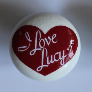 May include: Two white round knobs with a red heart design. The heart has the words "I Love Lucy" written in white cursive.