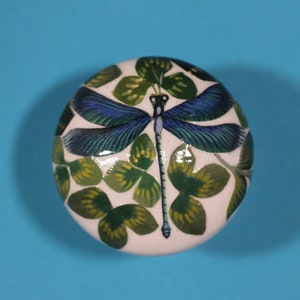 Peut inclure: Bouton de meuble rond et blanc avec un motif de libellule bleue et de feuilles vertes. Le bouton est posé sur un fond bleu vif. Un design inspiré de la nature pour la décoration.