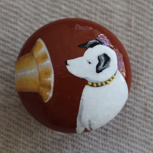 May include: A hand-painted rock with a brown background, a yellow and white hat, and a white dog with black ears and a gold collar.