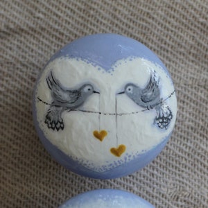 Może przedstawiać: Dwa pomalowane kamienie z ptakami. Pierwszy kamień jest jasnoniebieski z białym sercem i dwoma ptakami siedzącymi na drucie z wiszącymi sercami. Drugi kamień jest biały z trzema ptakami siedzącymi na drucie.