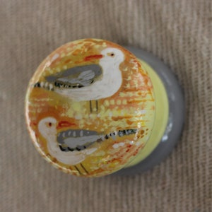 Pode incluir: Um pequeno prato de cerâmica com fundo amarelo e laranja e duas gaivotas pintadas. As gaivotas são brancas com asas cinzas e olhos pretos.