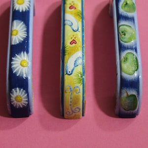 Könnte beinhalten: Drei dekorative Schrankknöpfe mit handbemalten Designs. Der erste Knopf hat einen blauen Hintergrund mit weißen Gänseblümchen. Der zweite Knopf hat einen gelben Hintergrund mit blauen Vögeln und roten Herzen. Der dritte Knopf hat einen blauen Hintergrund mit grünen Äpfeln.
