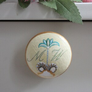 May include: A round, cream-colored drawer pull with a gold edge. The pull features a teal and cream floral design with the letters "M W" in a script font. Pink flowers and green leaves are in the background.
