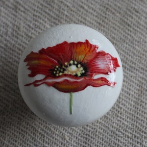 Könnte beinhalten: Ein runder weißer Knopf mit einer handbemalten roten Mohnblume. Die Blume hat einen gelben Mittelpunkt und einen grünen Stiel.