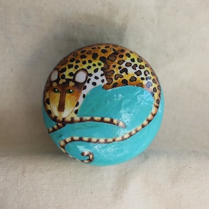 Op de afbeelding: Een handbeschilderde, ronde steen met een gestileerd luipaardontwerp. Het lichaam van het luipaard is beschilderd met gele en zwarte vlekken, terwijl het onderste deel een levendig turquoise is. Het gezicht van het luipaard is gedetailleerd met bruin en groen.