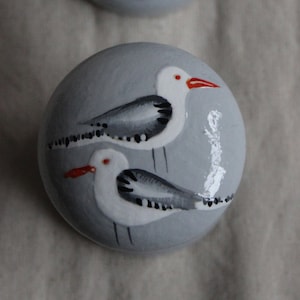 Puede incluir: Dos cajas redondas de madera pintadas de gris con tapas. Cada caja presenta una gaviota pintada a mano con marcas negras y blancas y un pico rojo.