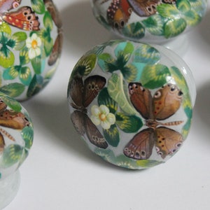 May include: A collection of ceramic cabinet knobs with a butterfly and floral design. The knobs feature a white base with a rounded top, adorned with detailed illustrations of butterflies, green leaves, and small white flowers. The butterflies have brown, orange, and red wings.
