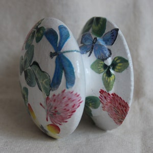 Könnte beinhalten: Zwei weiße, ovale Dekorationsgegenstände, möglicherweise eiförmig, werden gezeigt. Jedes ist mit handgemalten Blumen- und Insektenmotiven verziert. Eines zeigt eine blaue Libelle, das andere einen blauen Schmetterling und rosa Blumen.