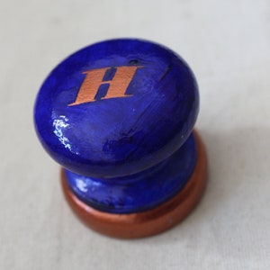 May include: A blue ceramic knob with a copper base and the letter "H" painted in copper on the top.