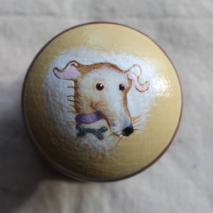 Op de afbeelding: Een ronde, crèmekleurige keramische sieradenbox met een geschilderd beeld van een hond die een paarse halsband en een bot draagt. De hond heeft witte vacht en roze oren.