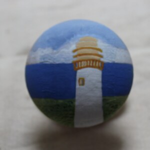 May include: A hand-painted, round decorative object featuring a lighthouse scene. The top portion depicts a blue sky with white clouds, transitioning to a blue sea. The lighthouse is white with a tan top and sits on a green base.