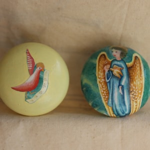 Puede incluir: Dos adornos redondos pintados a mano. Un adorno es amarillo con un pájaro rojo y una cinta azul. El otro adorno es verde con un ángel dorado que sostiene una paloma blanca.