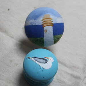 May include: Two hand-painted wooden knobs. One knob features a lighthouse scene with a blue sky and sea. The other knob is light blue with a painted seagull. These decorative knobs are perfect for cabinets or drawers.