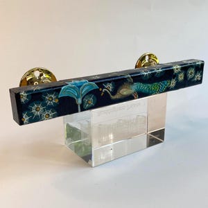 May include: A decorative cabinet pull with a rectangular black bar featuring a peacock and floral design. The pull is supported by a clear, rectangular base and has gold-colored circular mounting hardware.