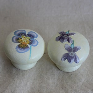 May include: Two white ceramic knobs with painted purple and blue flowers. The knobs are round and have a smooth surface.