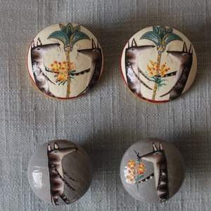 Pode incluir: Quatro puxadores decorativos redondos pintados à mão. Dois puxadores apresentam um fundo creme com uma palmeira central e figuras de animais. Os outros dois puxadores são cinzentos com uma única figura de animal e flores.