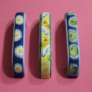 Op de afbeelding: Drie decoratieve kastknoppen met handgeschilderde ontwerpen. De eerste knop heeft een blauwe achtergrond met witte madeliefjes. De tweede knop heeft een gele achtergrond met blauwe vogels en rode harten. De derde knop heeft een blauwe achtergrond met groene appels.