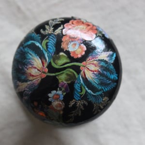 Puede incluir: Una caja redonda negra para joyas con un diseño floral. La caja está decorada con flores azules, rosas y amarillas sobre un fondo negro.