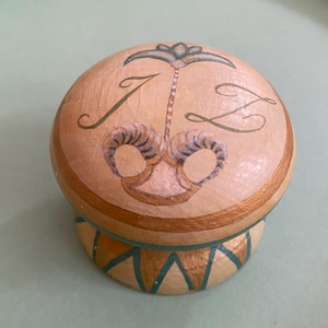 Könnte beinhalten: Kleine, runde, handbemalte Holzbox mit cremefarbenem Deckel und Boden. Der Deckel zeigt die Buchstaben "J L" und ein dekoratives Design in Gold und Türkis. Der Boden hat ein goldenes Band mit türkisfarbenen Dreiecken. Die Box steht auf einer hellgrünen Oberfläche.