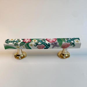 Könnte beinhalten: Ein weißer Schrankgriff mit floralem Design mit rosa Blüten, grünen Blättern und türkisfarbenen Akzenten. Der Griff wird von zwei goldfarbenen, abgerundeten Sockeln getragen.