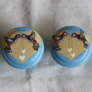 Könnte beinhalten: Zwei blaue und gelbe herzförmige Knöpfe mit gemalten Vögeln und weißen Herzen. Die Vögel schauen sich an und scheinen eine Schnur mit daran hängenden Herzen zu halten.